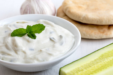 Tzatziki in white ceramic bowl with mint leaf garnish next to pita bread, cucumber and garlic bulb.