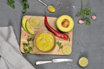 Mexican sauce guacamole in a bowl. Avocado, hot red pepper, slices of lime, garlic, salt lie on a wooden board. In the frame a knife and a napkin. Dark background. Close-up. View from above.