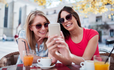 Friends meeting in a cafe. Young women drinking coffee and have fun with mobile phone. Consumerism, lifestyle concept