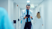 Determined Handsome Doctor Uses Digital Tablet Computer While Walking Through Hospital Hallway. Modern Bright Clinic With Professional Staff.