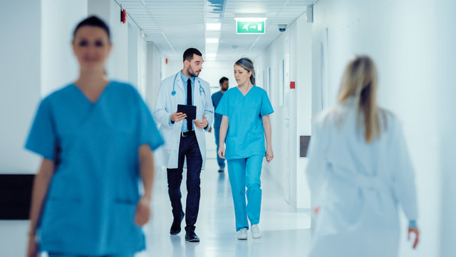 surgeon and female doctor walk through hospital hallway, they consult digital tablet computer while 