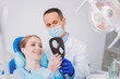 © Viacheslav Yakobchuk - Perfect result. Cheerful young woman sitting in a dental clinic and looking at the mirror while admiring her new beautiful braces