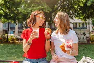 Two young female friends laugh and eat yoghurt outdoors