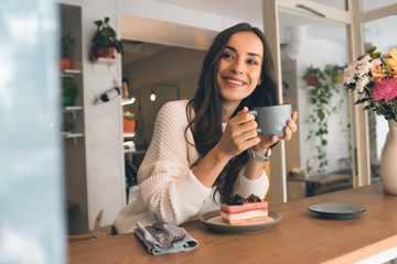 happy young woman with coffee cup sitting at table with cheesecake in cafe