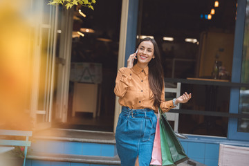 selective focus of happy stylish young woman with shopping bags talking on smartphone at city street