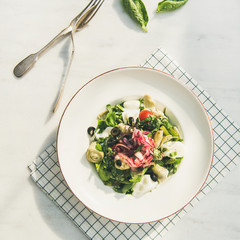 Wall Mural - Flat-lay of fresh green summer salad with artichokes, olives, soft cheese and red onion in white plate over light marble table background, top view, square crop