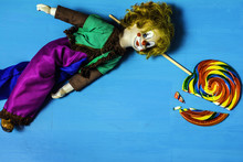 Antique Clown Doll With Blue Glass Eyes Red Nose And Orange Hair Laying On Wood Table Next To Large Broken Colorful Striped Lollipop On Blue Background
