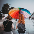 Two Girls Walking Across Square In Gloomy Rainy Weather Under Bright Umbrella Colored Like Rainbow. Concept Of Optimism And Love Of Life, Solving Any Problems And Difficulties. Instargam Style, Toned.