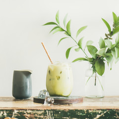 Wall Mural - Iced matcha latte drink in glass with coconut milk pouring from pitcher by woman's hand, white wall and plant branches at background, square crop. Summer refreshing beverage cold drink