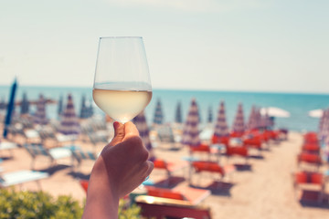 Female hand with a glass of white wine on the background of a beautiful beach. Travel vacations concept. Toned