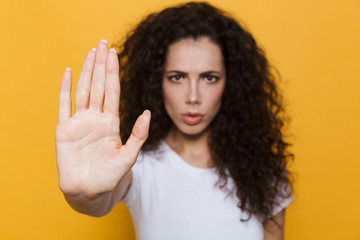 Wall Mural - Image of outraged woman 20s with curly hair doing stop gesture with hand, isolated over yellow background