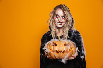 Poster - European witch woman wearing black costume and halloween makeup holding carved pumpkin, isolated over yellow background