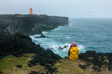 Rear View Of Woman In Yellow Raincoat Sitting On Cliff In Front Of Ocean And Looking At Lighthouse, Iceland