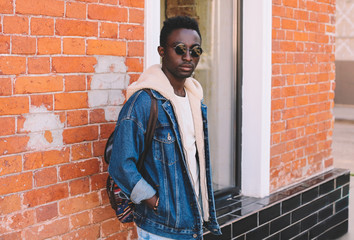 Fashion african man wearing jeans jacket, backpack poses on city street, brick wall background