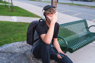 Wall Mural - Ashamed teenager sitting on a rock outside of a school while listening to music.