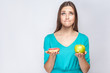 © khosrork - Portrait of dreaming young beautiful girl in blue blouse standing, holding pink donut and green apple in the hands like weighing with thoughful face on grey background. Indoor, isolated,copy space