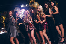 Cheers Girls! Beautiful Young Women In Evening Gown Holding Champagne Glasses And Looking At Camera With Smile While Celebrating In Nightclub