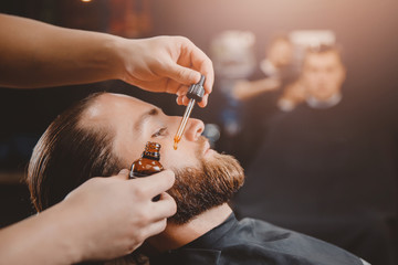 Barber applies beard oil with dropper for man in barbarshop.