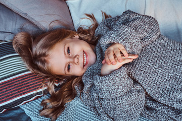 Close-up portrait of a little girl in warm sweater lying on bed.