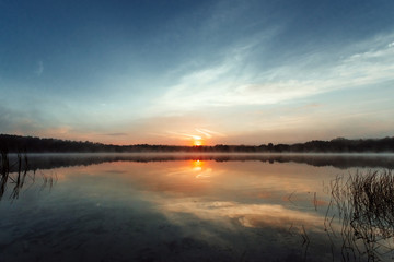 Beautiful, red dawn on the lake. The rays of the sun through the fog. The blue sky over the lake, the morning comes, the sky is reflected in the water.