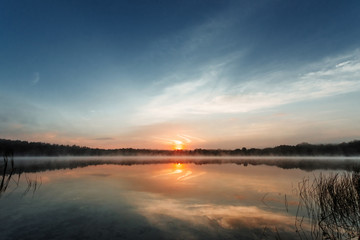 Beautiful, red dawn on the lake. The rays of the sun through the fog. The blue sky over the lake, the morning comes, the sky is reflected in the water.