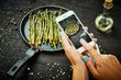 woman's hands are holding the phone and taking a picture of delicious young asparagus on a wooden table