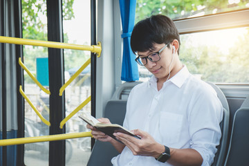 Young Asian man traveler sitting on a bus listening music and reading book while smile of happy, transport, tourism and road trip concept