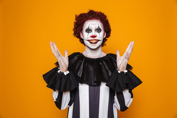 Young clown man 20s wearing black costume and halloween makeup looking at camera, isolated over yellow background