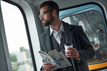 Business trip. Young businessman standing in the train holding newspaper and cup drinking hot coffee enjoying the window view close-up