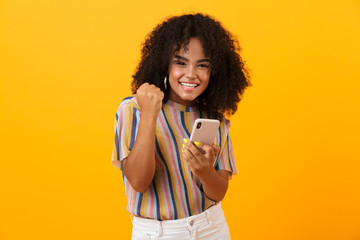 emotional african woman posing isolated over yellow background using mobile phone make winner gestur