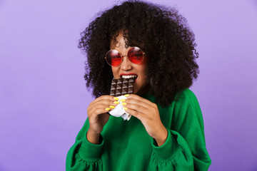 Poster - Excited pretty african woman isolated over violet background eat chocolate.