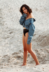 Portrait of young beautiful woman posing on sand. Model wearing stylish blue denim jacket and black panties. Sunset light. Female fashion concept.