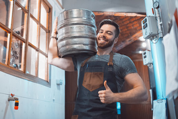 Wall Mural - A young brewer in an apron holds a barrel with beer in the hands of a brewery