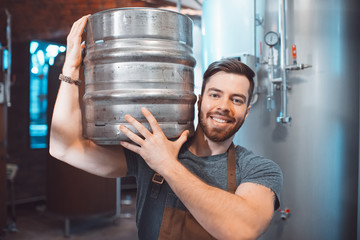 A young brewer in an apron holds a barrel with beer in the hands of a brewery