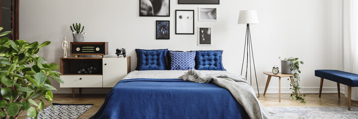 Real photo of an eclectic bedroom interior with graphics on a wall, blue pillows and radio on a cupboard