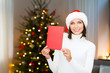 © Syda Productions - holidays, celebration and people concept - smiling woman in santa hat holding greeting card over christmas tree lights background