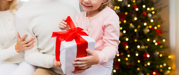 family, holidays and people concept - close up of happy mother, father and little daughter with gift box sitting on sofa at home over christmas tree lights background