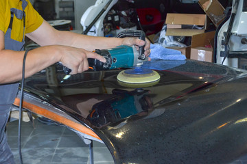 The master man of the detailing in work clothes and dirty hands polishes the bodywork of the bonnet of the car in black with a polishing machine in the workshop for body repair cars