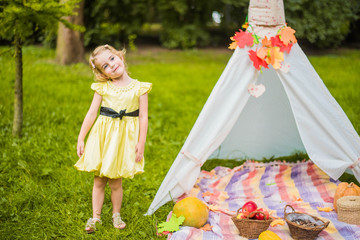 Little girl lying and playing in a tent, children's house wigwam in park Autumn portrait of cute curly girl. Harvest or Thanksgiving. autumn decor, party, picnic. Child in yellow dress with apple 