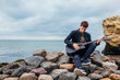 © maryviolet - Young man with acoustic guitar playing on beach surrounded with rocks on rainy day
