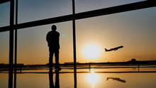Silhouette Of A Tourist Guy Watching The Take-off Of The Plane Standing At The Airport Window At Sunset In The Evening. Travel Concept, People In The Airport.