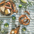Flat-lay of summer picnic set with fruit, cheese, sausage, bagels and lemonade over striped blanket, top view, square crop
