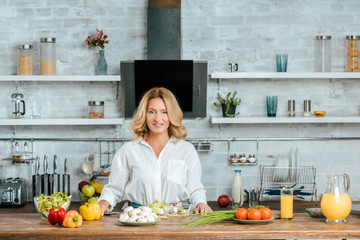 happy adult woman with various fresh vegetables on kitchen table looking at camera