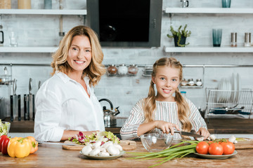 Wall Mural - smiling adult mother and little daughter cooking together and looking at camera