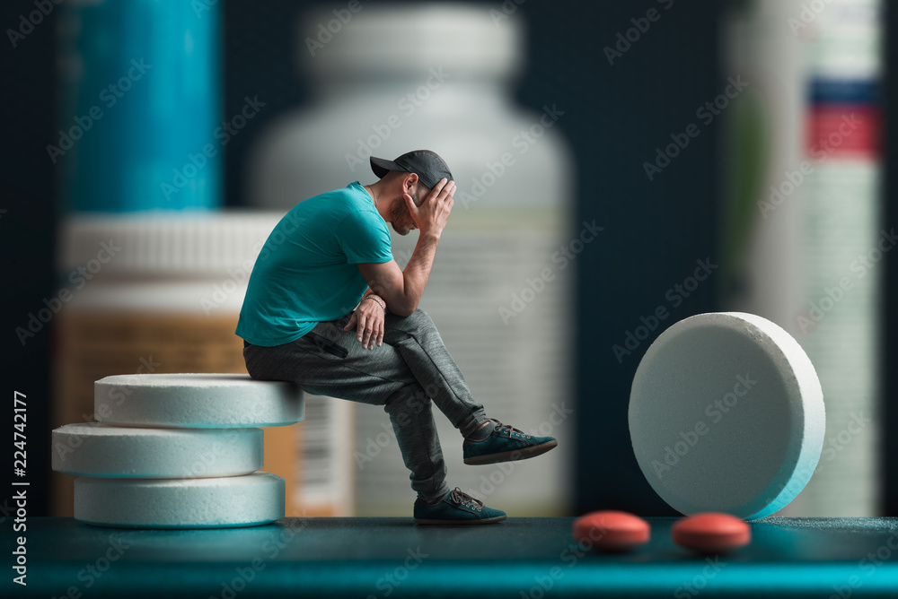 The man sitting on the pills. He's depressed. Flacons of medicines in the background - obrazy, fototapety, plakaty 
