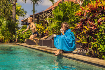 Sick man traveler. The man caught a cold on vacation, sits sad at the pool drinking tea and blows his nose into a napkin. His son is healthy and swimming in the pool. Travel insurance concept