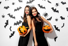 Two Beautiful Brunette Women In Black Dresses Have Fun With Jack-o-lanterns On A White Background With Bats And Spiders