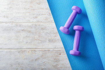 Yoga mat with dumbbells on light wooden background