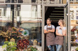 © LIGHTFIELD STUDIOS - cheerful caucasian florists standing near flower shop and looking away