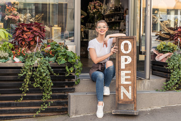 Wall Mural - attractive smiling flower shop owner sitting on stairs with open signboard